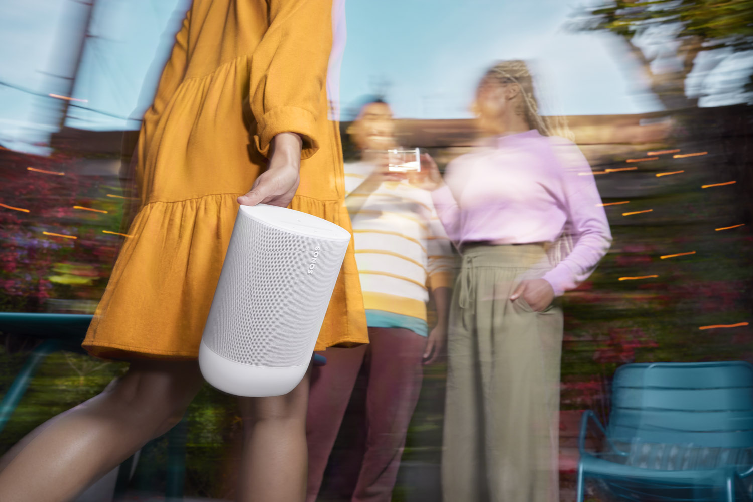 En person i en orange klänning håller i en Sonos Move 2 högtalare utomhus. I bakgrunden suddiga gestalter som verkar njuta av sällskap och musik. Högtalarens design är modern och anpassad för utomhusbruk med sitt IP56-klassade skydd mot damm och fukt. Scenen frammanar en känsla av frihet och njutning, med en atmosfär av kontinuerlig underhållning som kan pågå upp till 24 timmar.