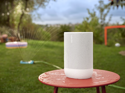 Sonos Move 2 högtalaren står centralt placerad på ett rött bord utomhus, omgiven av en trädgårdsmiljö med en vattenspridare i funktion och leksaker i bakgrunden. Högtalaren är täckt av vattendroppar, vilket visar dess fukttålighet. Det robusta utseendet på Move 2 accentueras av dess våta yta, vilket understryker dess lämplighet för användning i olika utomhusaktiviteter och dess IP56-klassning.