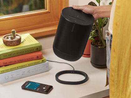 En person lyfter en svart Sonos Move 2 högtalare från dess laddningsring på ett köksbord. Intill högtalaren syns en smartphone och böcker om matlagning, vilket antyder en avkopplande eller inspirerande miljö. Växter och ett fönster som släpper in naturligt ljus ger en hemtrevlig och mysig atmosfär. Högtalaren är designad för att enkelt kunna bära med sig och ladda på ett enkelt sätt, vilket möjliggör en smidig integration i vardagslivet.