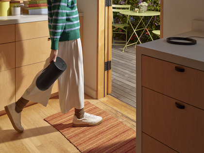 En person i en vita byxor, grön- och vitrandig tröja och vita skor går genom en ljust inredd lägenhet medan de bär en svart Sonos Move 2 högtalare. Dörren står öppen ut till en solig balkong, vilket förmedlar en känsla av att musiken och användaren rör sig från ett rum till ett annat. Inredningen är modern och minimalistisk, och högtalarens stilrena design smälter väl in i hemmamiljön samtidigt som den erbjuder bärbarhet och flexibilitet.
